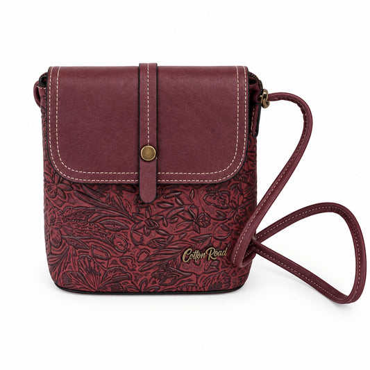 A burgundy leather crossbody bag with a tonal floral embossed pattern on the front flap, a gold-tone snap button, and a matching leather strap, resting on a white shelf.